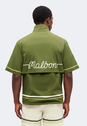 Mann trägt ein olivgrünes kurzärmeliges Shirt mit weißem "malbon" Text auf dem Rücken und weiße Shorts mit grünen Streifen, steht mit dem Rücken zur Kamera.