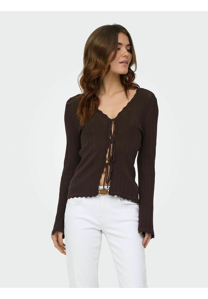 Femme aux cheveux bruns ondulés portant un cardigan côtelé marron foncé à manches longues noué devant et un pantalon blanc avec une ceinture.