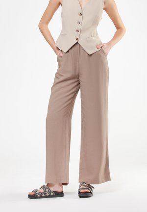 Donna che indossa un gilet beige senza maniche con bottoni, pantaloni larghi taupe morbidi e sandali slip-on con motivo, in piedi con le mani nelle tasche.