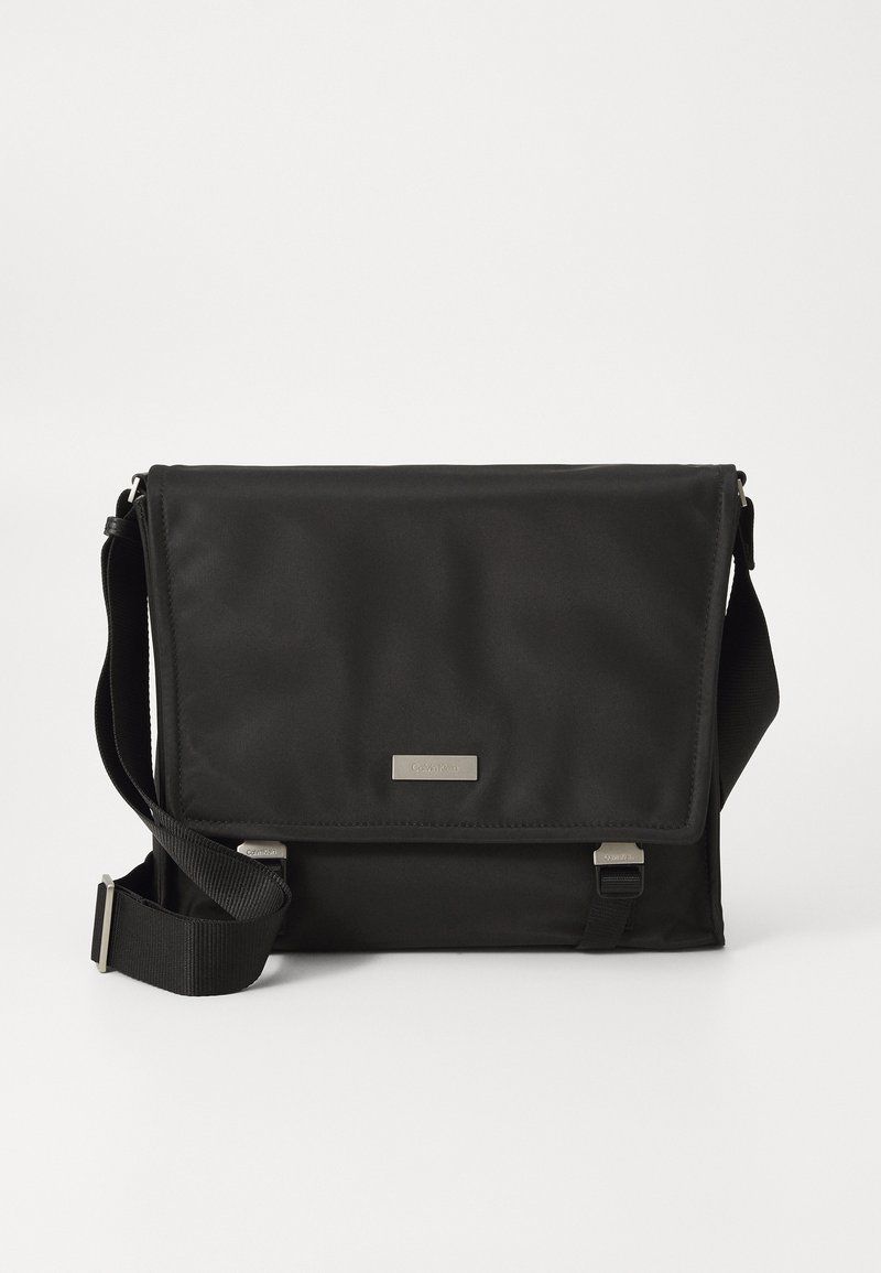 Sac messager noir en nylon avec une finition mate. Il dispose d'une fermeture à rabat, d'une sangle réglable et d'une petite plaque en métal avec logo.