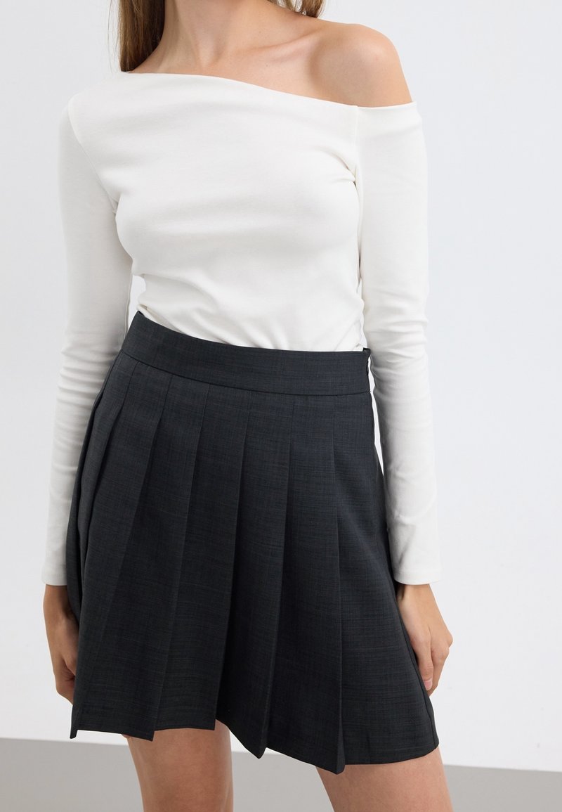 Femme portant un haut blanc à manches longues et épaules dénudées, associé à une jupe plissée noire taille haute, debout devant un fond uni.