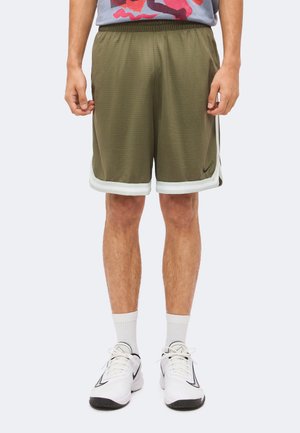 Man draagt olijfgroene basketbalshorts met witte bies, witte sokken tot halverwege de kuit en witte Nike sneakers met zwart Swoosh-logo.