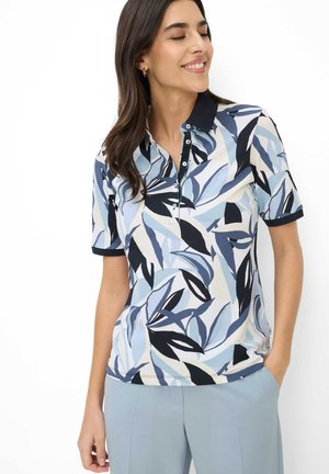 Donna sorridente con gli occhi chiusi che indossa una polo a maniche corte con motivi di foglie blu e nere e pantaloni azzurri, con la mano in tasca.