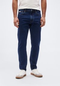 Σκούρα μπλε τζιν με ίσια γραμμή, πέντε τσέπες και μπροστινό κούμπωμα με κουμπιά. Συνδυασμένα με λευκά αθλητικά παπούτσια.