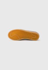 Semelle de sneaker avec un fond en caoutchouc beige présentant un motif en chevron, des bords lisses et une légère élévation pour un soutien en matière d'amorti.