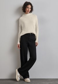 Crème ribgebreide trui met een hoge nek, gecombineerd met zwarte wijde pantalons en beige sneakers, tegen een effen grijze achtergrond.