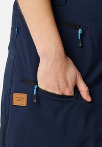 Marinekleurige stof met een geprofileerde afwerking, voorzien van een zichtbare zak met een blauwe ritssluiting en een bruin leren merkaanduiding.