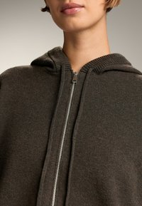 Pulover cu glugă, cu fermoar, de culoare maro închis, dotat cu guler ribbed, fermoar metalic și textură moale din tricot. Fără logo-uri sau modele vizibile.