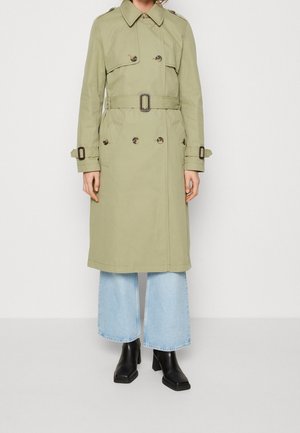 Kvinde iført en knælængde beige trenchcoat med bælte over lyseblå vidde bukser og sorte ankelstøvler med hæl.