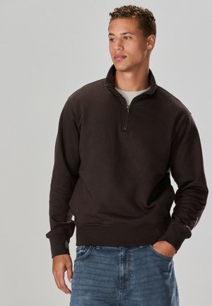 Ung mand med kort krøllet hår iført en mørkebrun kvart-lynlås sweatshirt og blå jeans, stående med den ene hånd i lommen.