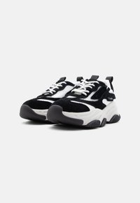 Χοντρά ασπρόμαυρα αθλητικά παπούτσια με πάνελ από δίχτυ και σουέτ, λευκά κορδόνια, παχιά υφή στη σόλα και θηλιές στο πίσω μέρος, σε ένα απλό φόντο.