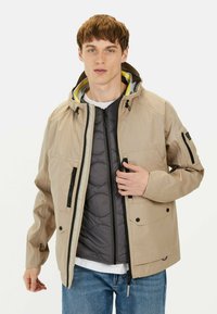 Lys beige vandtæt jakke med grå inderside, lynlåslommer, justerbar hætte og gule detaljer. Model i blå jeans.