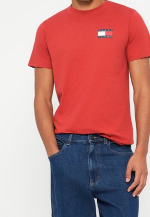 Mężczyzna ubrany w czerwoną koszulkę Tommy Jeans i niebieskie dżinsy, z jedną ręką w kieszeni, na jednolitym tle.