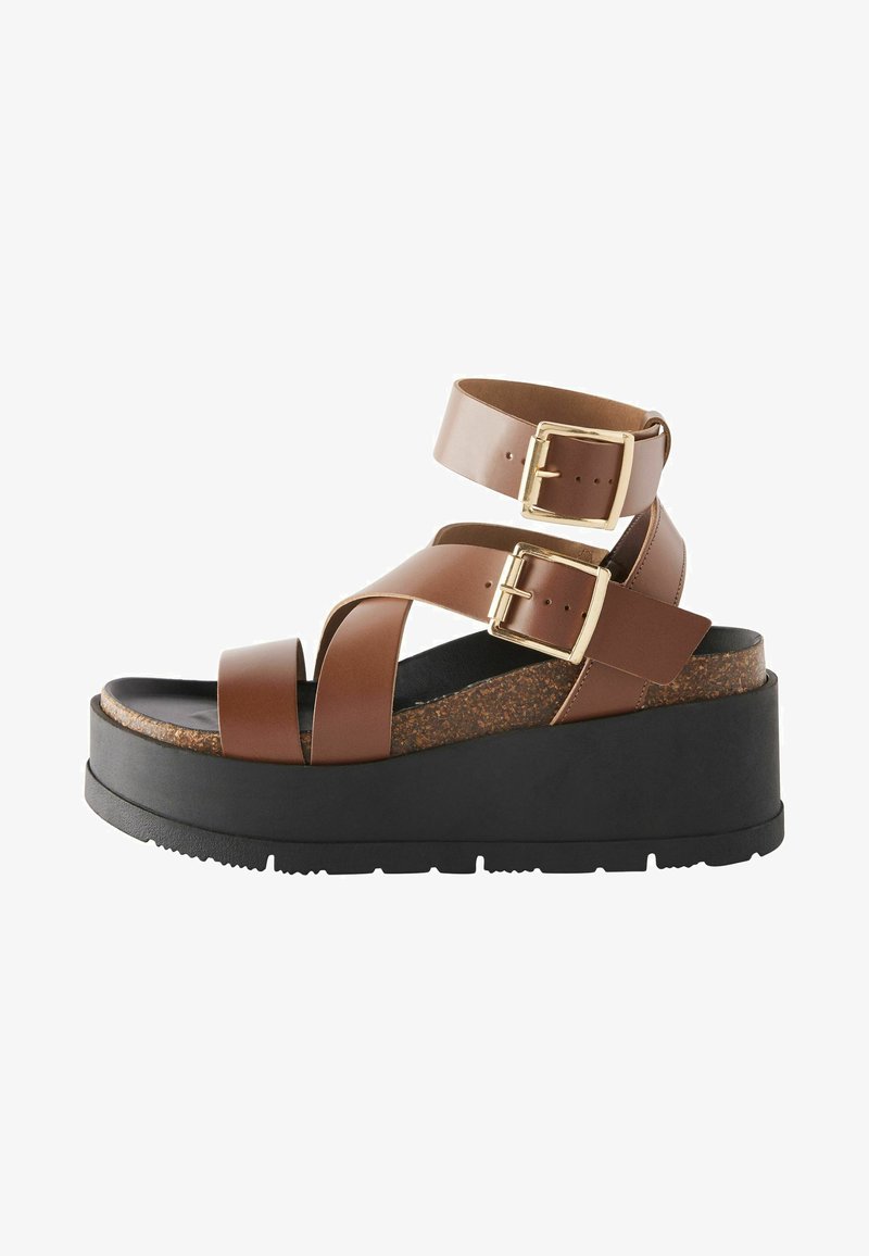 Brown platform sandals with adjustable buckle straps, cork footbed, and a textured black rubber sole. Two crossed straps across the foot.