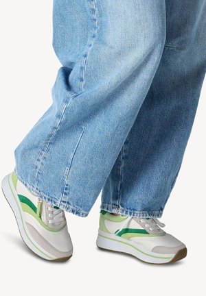 Sneakers with a white and grey upper, green accents, and a textured sole. Worn with loose blue denim pants, showcasing a casual style.