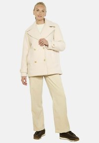 Beżowy dwurzędowy płaszcz z złotymi guzikami, szerokim kołnierzem i bocznymi kieszeniami, zestawiony z jasnymi, teksturowanymi spodniami i ciemnymi trampkami.