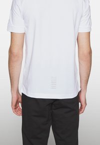 Camiseta blanca de manga corta, con un detalle rayado texturizado en la parte baja de la espalda, combinada con pantalones negros.