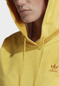 Gul hoodie i manchester med ett texturerat tyg, utrustad med en framträdande dragsko och broderad Adidas-logga på bröstet.