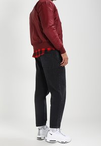 Röd bomberjacka med ribbade manschetter över en svart och röd rutiga skjorta, matchad med löst sittande svarta jeans och vita sneakers.
