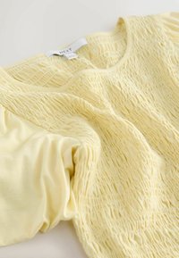 Sárga felső puha anyagból, gyűrt textúrával és kerek nyakkivágással. Rövid, puffos ujjakkal és "NEXT" feliratú címkével rendelkezik.