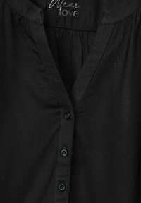 Blusa nera con scollatura a V, apertura frontale con sei bottoni e leggero luccichio. Il materiale è liscio con una lieve texture. "Indossa amore" stampato sul colletto.