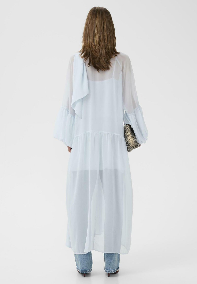 Femme portant une longue robe légère bleu clair transparente sur un jean et des talons, tenant une pochette texturée, vue de dos sur un fond uni.