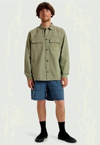 Cămașă verde cu nasturi, cu două buzunare pe piept, purtată peste pantaloni scurți albaștri. Prezintă un logo cu soare pe buzunarul stâng și pantofi negri cu șosete până la glezne.