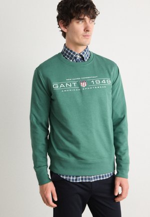 Grüner Pullover aus Baumwollmischung mit Rundhalsausschnitt, gerippten Bündchen und GANT-Logo auf der Brust, über einem karierten Hemd getragen.