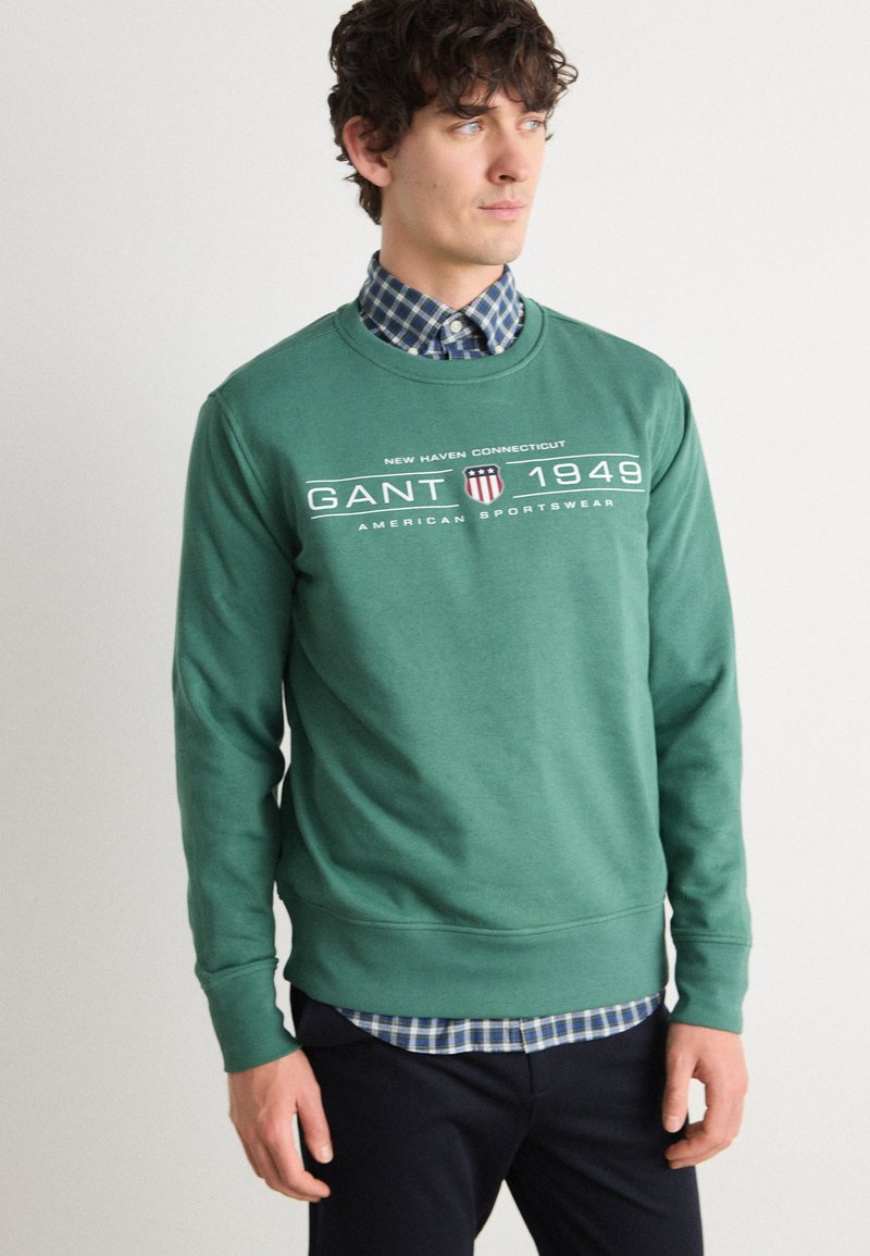 Felpa verde in misto cotone, con colletto rotondo, polsini a coste e dettaglio del logo GANT sul petto, indossata sopra una camicia a quadri.