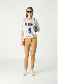 T-shirt branca com estampado gráfico azul, calças cargo slim-fit em tom areia, e ténis brancos. Os acessórios incluem uma pequena mala azul e óculos de sol.