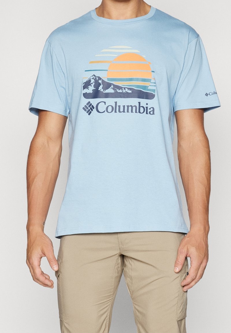 Camiseta de manga corta azul claro con gráfico de montaña y sol y logo de "Columbia", combinada con pantalones cargo beige.