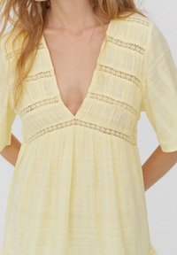 Gelbes Kleid mit tiefem V-Ausschnitt, kurzen Ärmeln und strukturiertem Stoff. Verfügt über Häkelverzierungen in horizontalen Streifen über dem Oberteil.