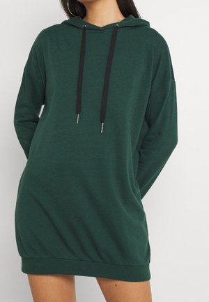 Femme portant une robe à capuche vert foncé avec des cordons noirs, debout devant un fond blanc uni.