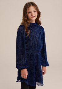 Mörkblå klänning med smock, långa puffiga ärmar, texturerat skimrande tyg, kjol i lager och en vågig nederkant. Buren över svarta strumpbyxor.