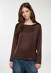 Maglietta marrone a maniche lunghe realizzata in tessuto morbido e lucido con una ampia scollatura a barca. Abbinata a jeans di denim blu larghi.