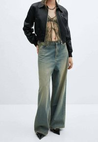 Personne portant une veste courte noire, un haut en dentelle olive translucide noué à l'avant, un jean large délavé et des chaussures noires pointues.