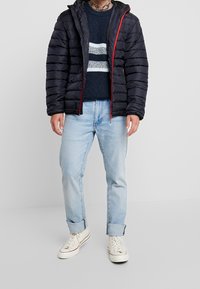 Schwarze Steppjacke mit roten Reißverschlussdetails, getragen über einem marineblauen Pullover mit weißen Streifen. Kombiniert mit hellblauen Jeans und beigefarbenen Sneakers.