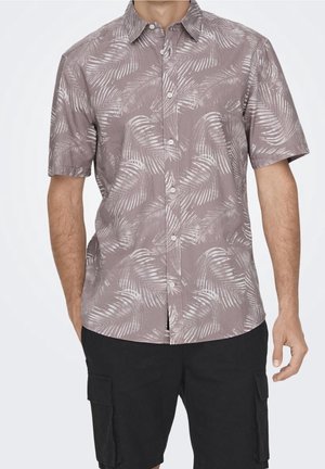 Mand iført kortærmet skjorte med lysegråt palmebladsmønster og sorte cargoshorts, stående med armene afslappet ned langs siden.