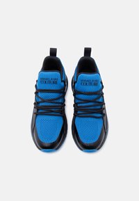 Μπλε και μαύρα αθλητικά παπούτσια με επάνω μέρος από δίχτυ, καουτσούκ σόλα και μαύρα κορδόνια. Το όνομα της μάρκας είναι τυπωμένο σε μια μπλε γλώσσα.