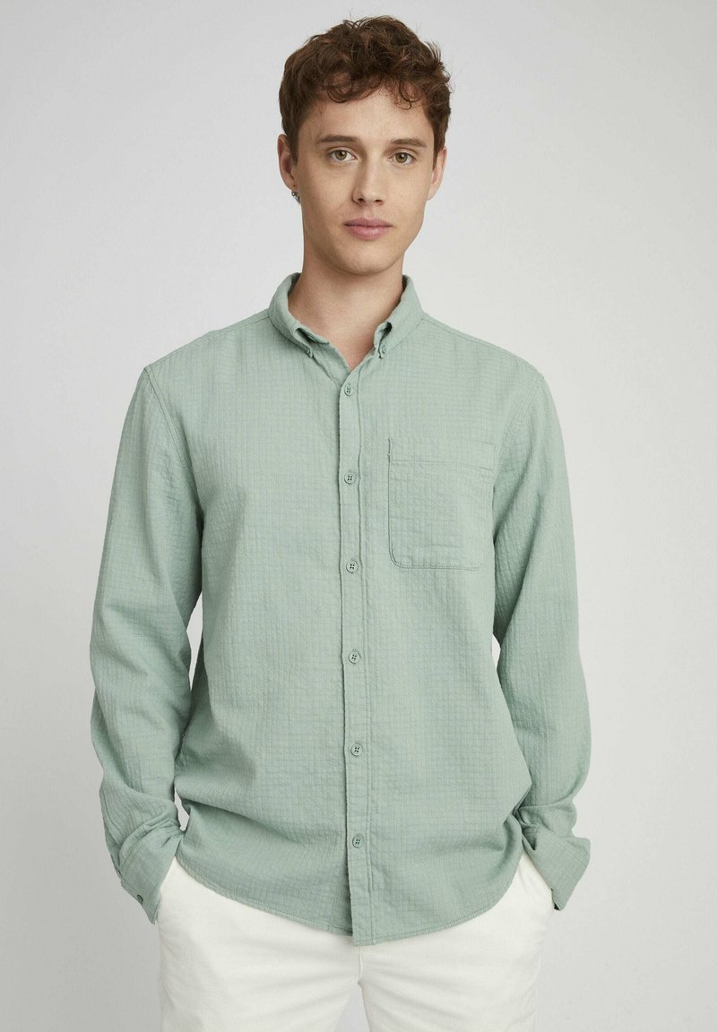 Chemise à carreaux verts avec une poche sur la poitrine, manches longues, en tissu doux, portée avec un pantalon de couleur claire.