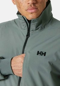 Chaqueta impermeable gris con alto cuello, cierre de cremallera, puños elásticos y logo negro en el pecho. Textura suave y diseño ajustado.