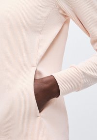 Χέρι με σκουρόχρωμο δέρμα που έχει τοποθετηθεί στην πλευρική τσέπη ενός ανοιχτό ροζ μακρυμάνικου πουκάμισου με υφή, με φόντο λευκό.