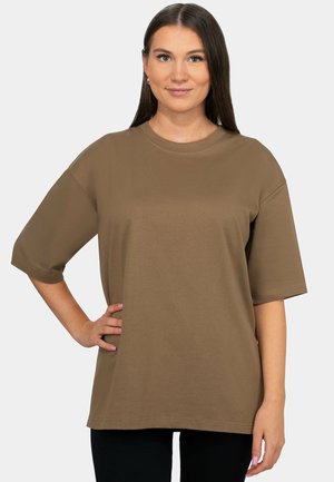 Young woman with long brown hair wearing a loose brown oversized T-shirt, posing with one hand on her hip, neutral background.