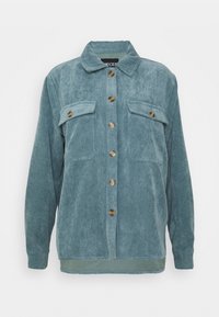 Camicia di corduroy in blu polveroso. Presenta una chiusura a bottoni, due tasche sul petto e un tessuto testurizzato con righe verticali.