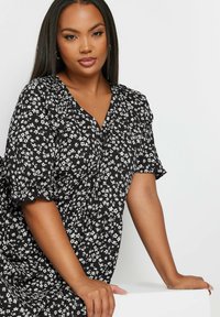 Musta kukallinen mekko, jossa on pieniä valkoisia kukkia, lyhyet puff-hihoja, V-pääntie ja kerroksellinen muotoilu. Pehmeä kangas, jossa on hieman tekstuuria.