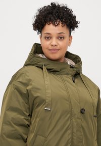 Tamsiai žalia izoliacija parka su kapišonu, užsegama zipu ir mygtukais, su šoninėmis kišenėmis ir reguliuojamomis raištelėmis. Lygus paviršius.