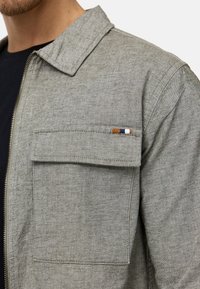 Col et poche poitrine d'une veste zippée grise portée sur une chemise noire, avec une petite étiquette à rayures marron, bleu marine et blanche.
