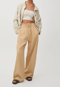 Pantalon beige surdimensionné avec une taille à cordon, des plis et deux boutons à l'avant. Associé à un haut court blanc en maille côtelée et une veste légère.