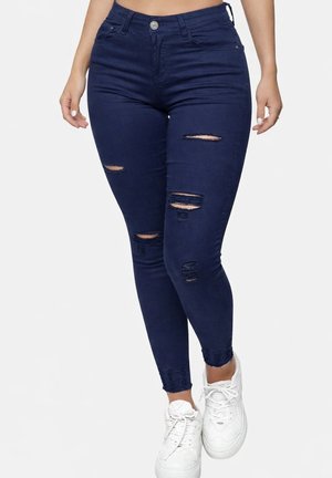 Kvinde iført mørkeblå, revnede skinny jeans og hvide sneakers, stående med hænderne afslappet ned langs siden.