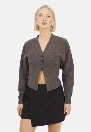 Donna bionda che indossa un cardigan corto marrone con bottoni e una minigonna asimmetrica nera, in piedi davanti a uno sfondo semplice.